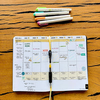Open 2026 planner with daily schedule on a wooden surface, surrounded by pens.